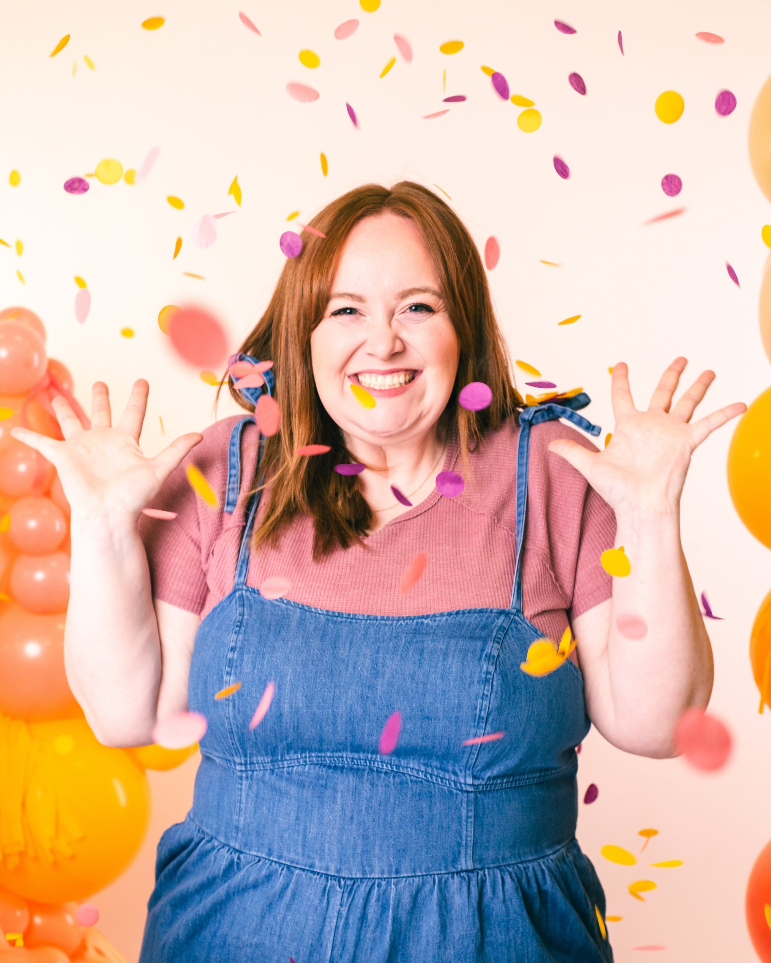 Woman in denim overalls and pink shirt with confetti and balloons on a light pink background