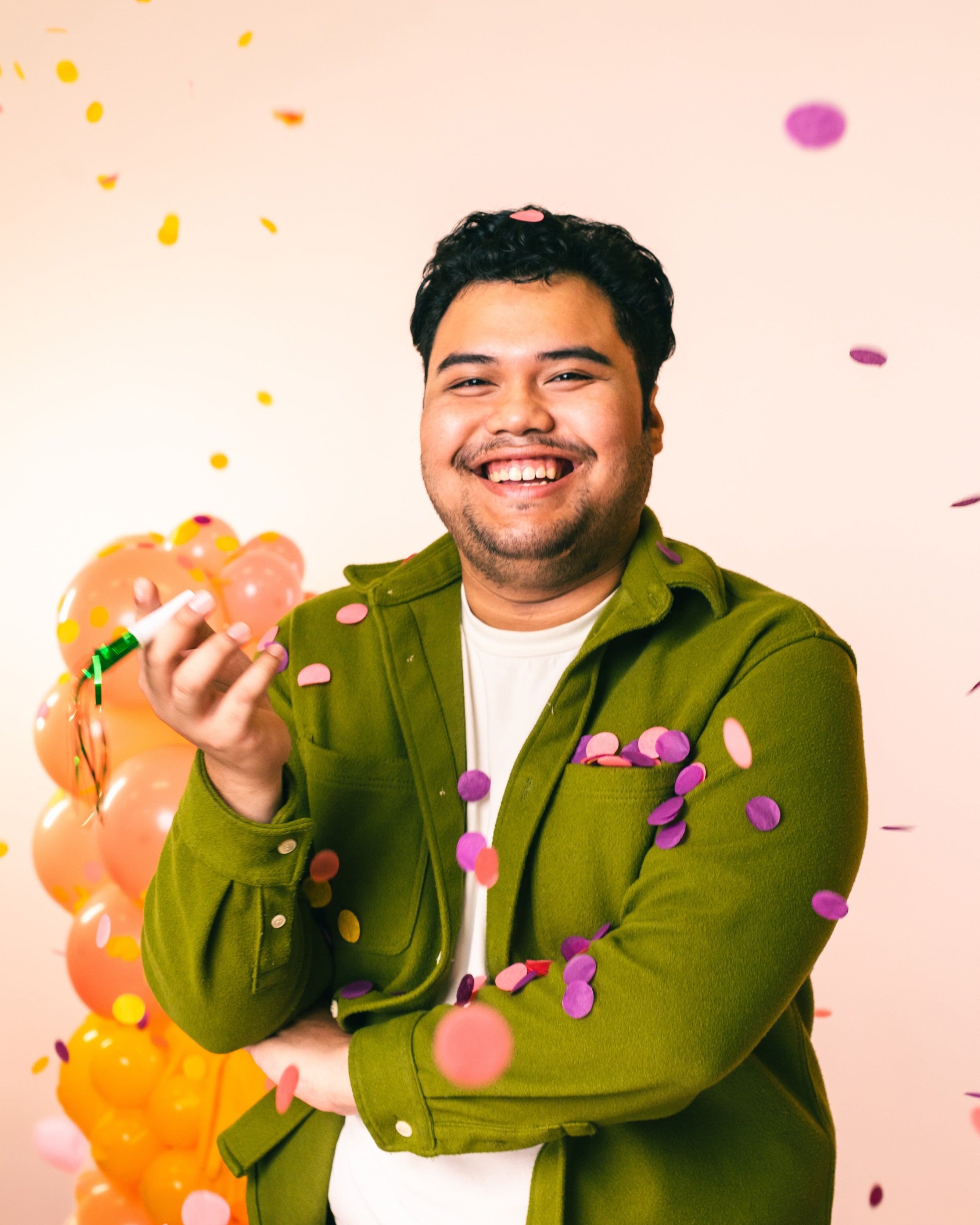 Man in a green jacket holding a noisemaker with confetti around him smiling at the camera on a light pink background