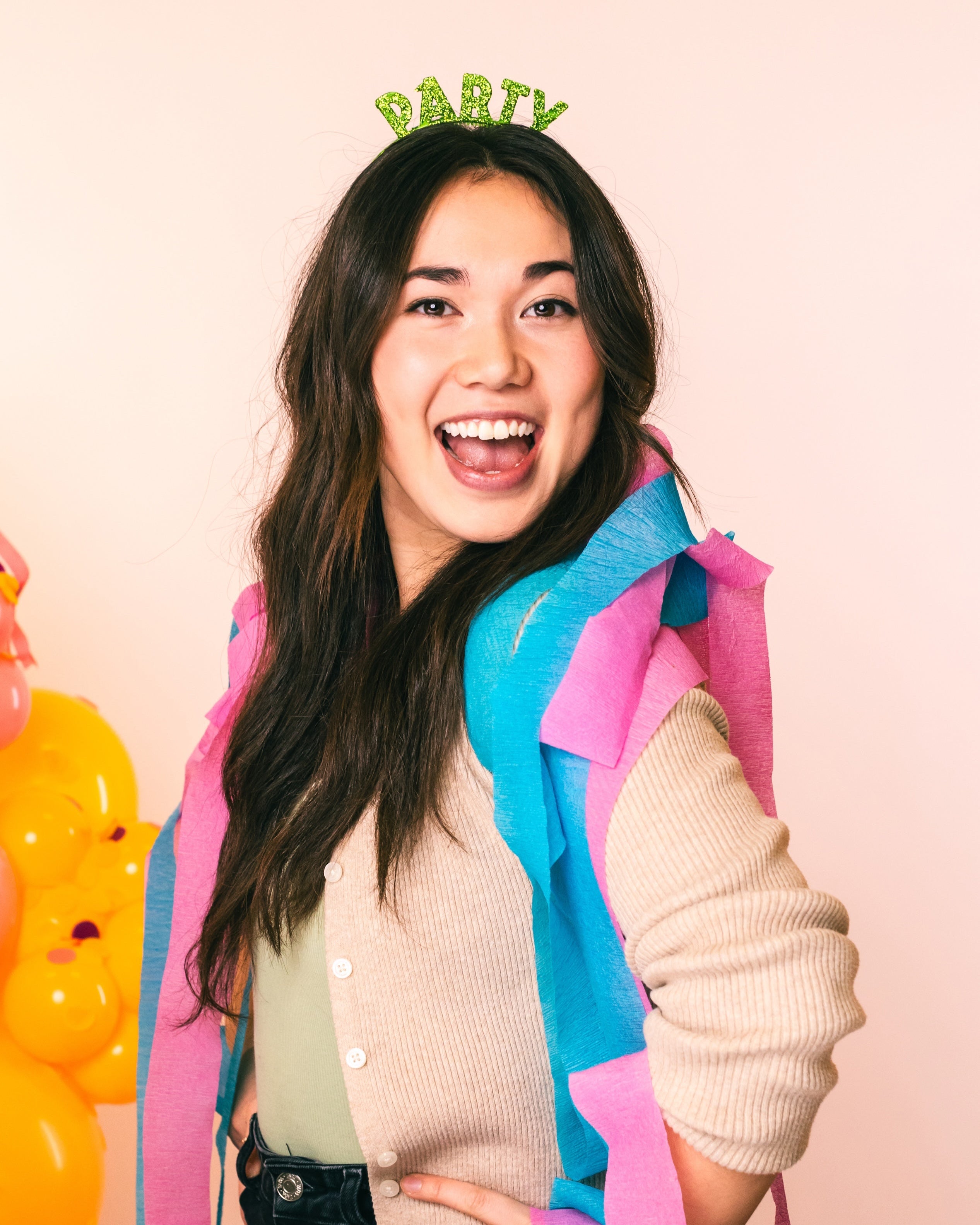 Woman wearing party streamers as a scarf and a headband that reads "party" smiling at the camera on a light pink background