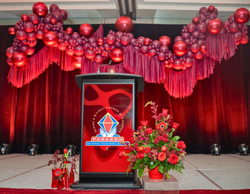 Paragon Awards podium. The balloon and fringe backdrop is bottom lit by red lights. The backdrop is chrome red orbs and burgundy balloons with red fringe below. 