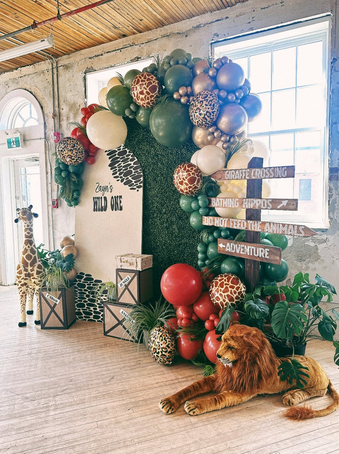 A full shot of our safari balloon install. It features a stuffed lion and giraffe, a balloon garland with the colors red, white, green, and beige; animal printed balloons, and a grassy backdrop. A white backboard reads: Zayn's wildone.