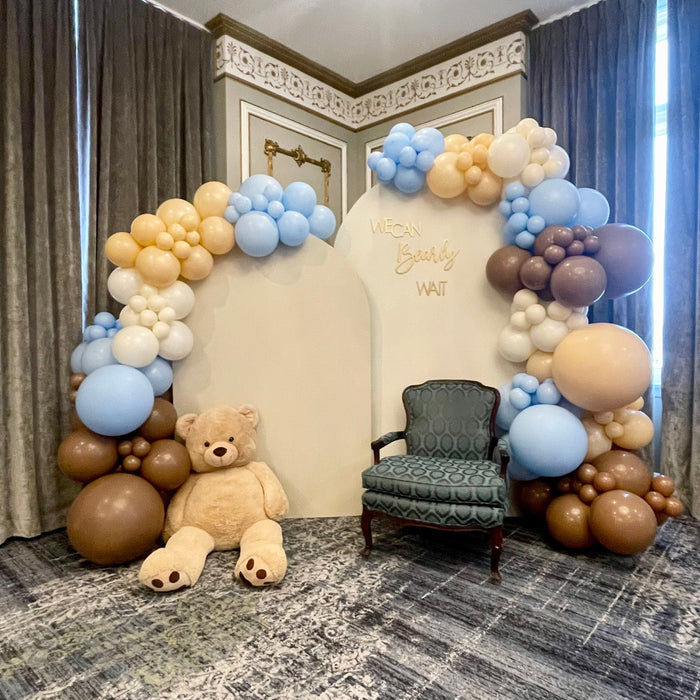 A cute backdrop featuring a giant teddy bear and a balloon garland with the colors baby blue, beige, brown, and creamy white. It reads 
