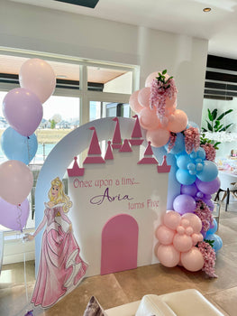 A castle backdrop is decorated with a pink, purple, and blue balloon garland. Pink flowers are clustered between the balloons. A sleeping beauty stand holds some more balloons in front of the castle. The backdrop reads: once upon a time...Aria turns five.