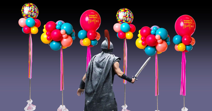 A man with a sword and knight armour stands in with his back facing the audience in front of a bunch of balloons