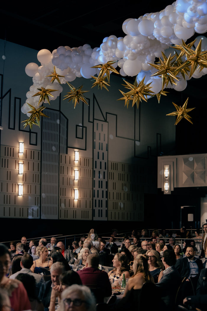 Habitat for Humanity's Home by Midnight Gala; with golden foil stars and white balloon clouds. People in the audience smile and talk together.