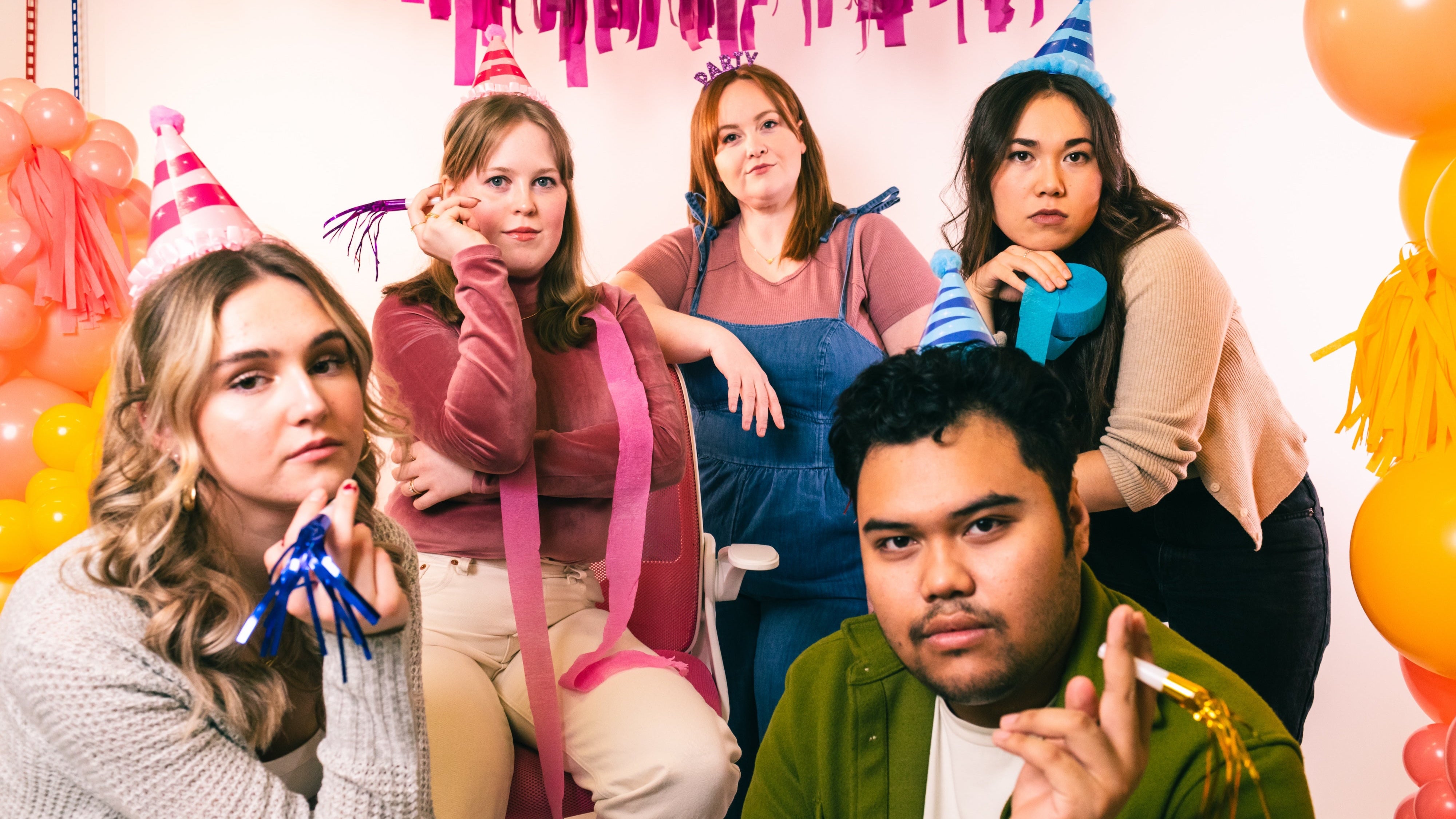 Group of people with serious facial expressions wearing party hats and surrounded by balloons and streamers 